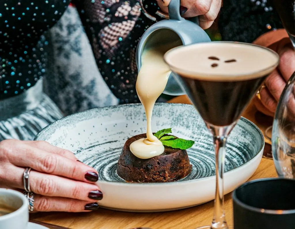 Someone pouting custard on their Christmas Pudding. There is a cocktail in front of them